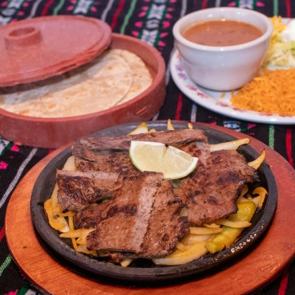 Sizzling steak fajitas with onions and a lime wedge, alongside tortillas, beans, rice, and salad.