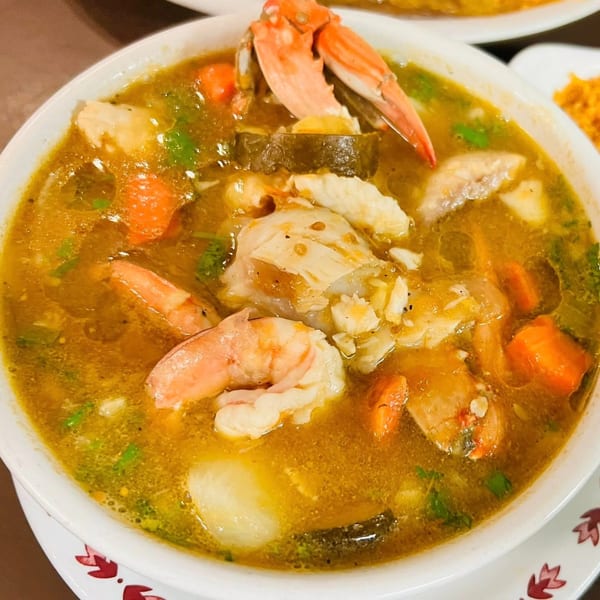 A close-up of a vibrant seafood soup with crab, shrimp, and vegetables in a white bowl.